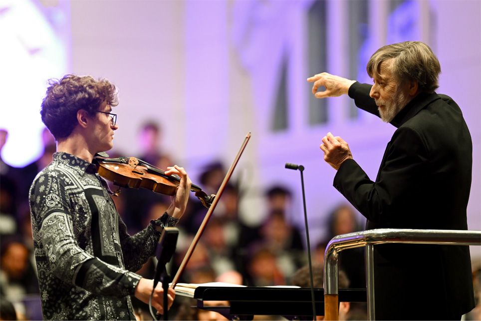 A male violinist, wearing a smart shirt, looking at a male conductor with grey hair and a beard, wearing smart black attire, who is conducting an orchestra with his arms.